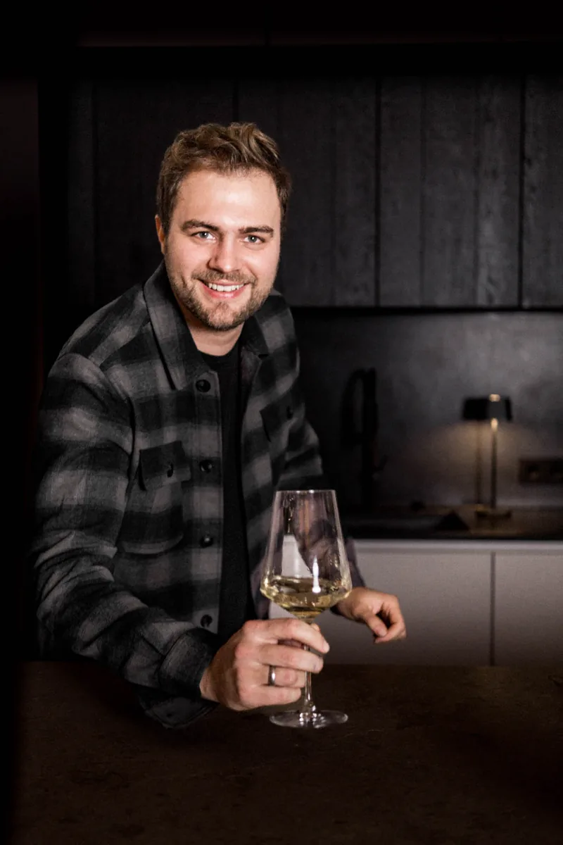 Jonas von 7 Zutaten lächelt an einer Küchentheke und hebt ein Glas Weißwein